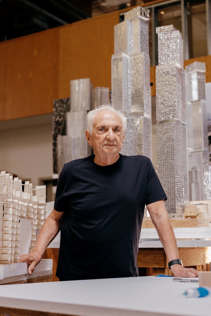 Frank Gehry wearing a black shirt and standing in front of a silver-colored model of a city skyline.