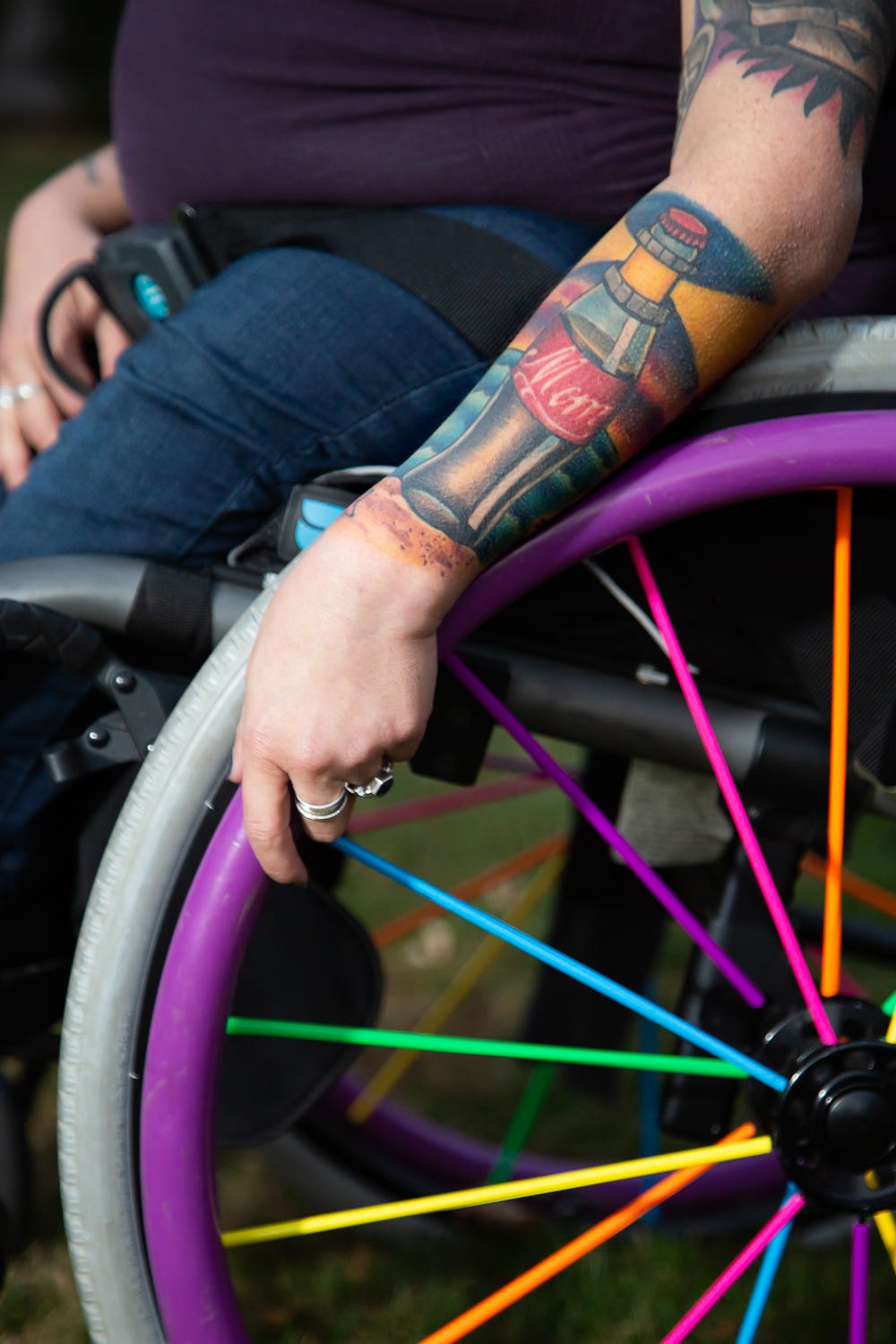 A close-up shot of Valarie’s forearm tattoo. The tattoo is of a Coca-Cola bottle on the beach that has “Mom” written on the label. Her hand has two rings on it and is holding the purple push rim of her wheelchair. The rainbow spoke covers are visible in the lower right hand corner of the picture.