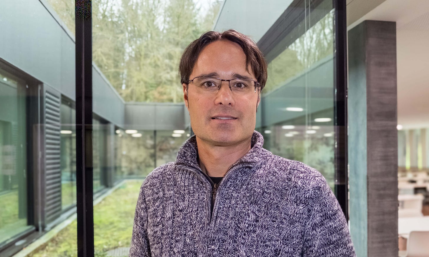 Das Foto zeigt den Psychologen Thomas Brunner, Professor für Konsumenten-Verhalten an der BFH-HAFL in Bern. Er trägt eine Brille und einen grau melierten Pullover mit Reissverschluss. Im Hintergrund ist durch grosse Glasfenster ein Innenhof mit grünem Gras und dahinterliegendem Wald sichtbar. Die moderne Architektur des Gebäudes mit klaren Linien und Glasflächen unterstreicht die professionelle Umgebung.