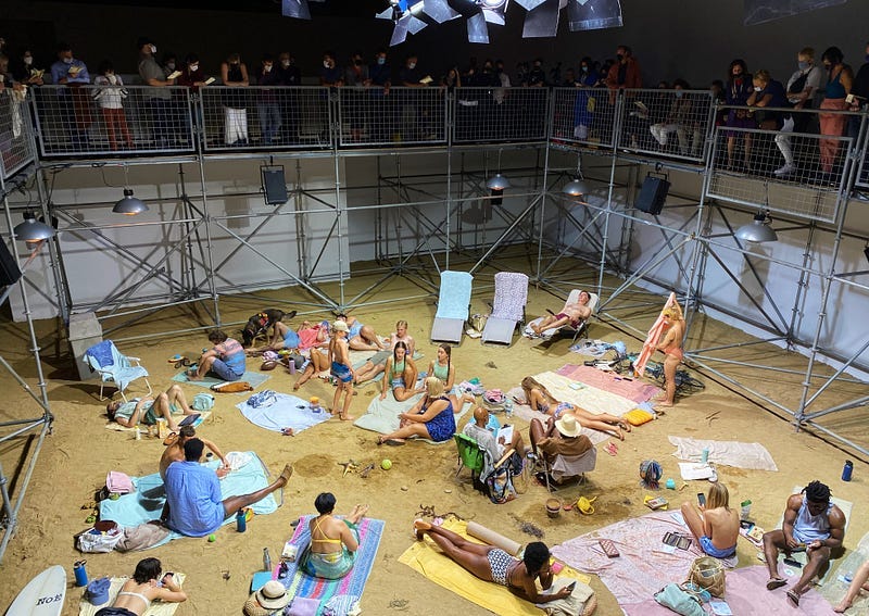 The setting is a fake beach. It’s inside a museum’s gallery. The “beach” is comprised of sand and performers are dressed in bathing suits, wetsuits, and casual summer garb. They lounge about on their towels or beach chairs, snacking, reading, and chatting. Surrounding them is metal scaffolding. Masked museum goers line the edges of the scaffolding, peering down on the scene.