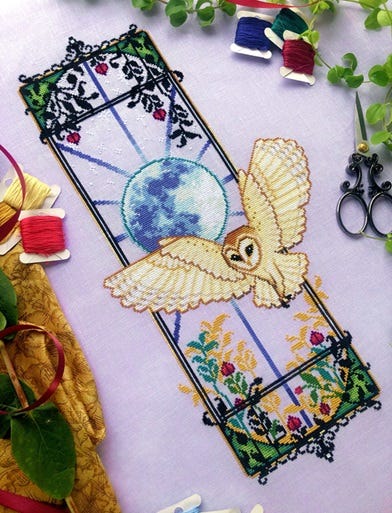 A cross stitch of a white barn owl, flying with wings extended.  An ornate art nouveau-style window is behind the owl, with foliage details.  A bright blue full moon glows from behind the owl.