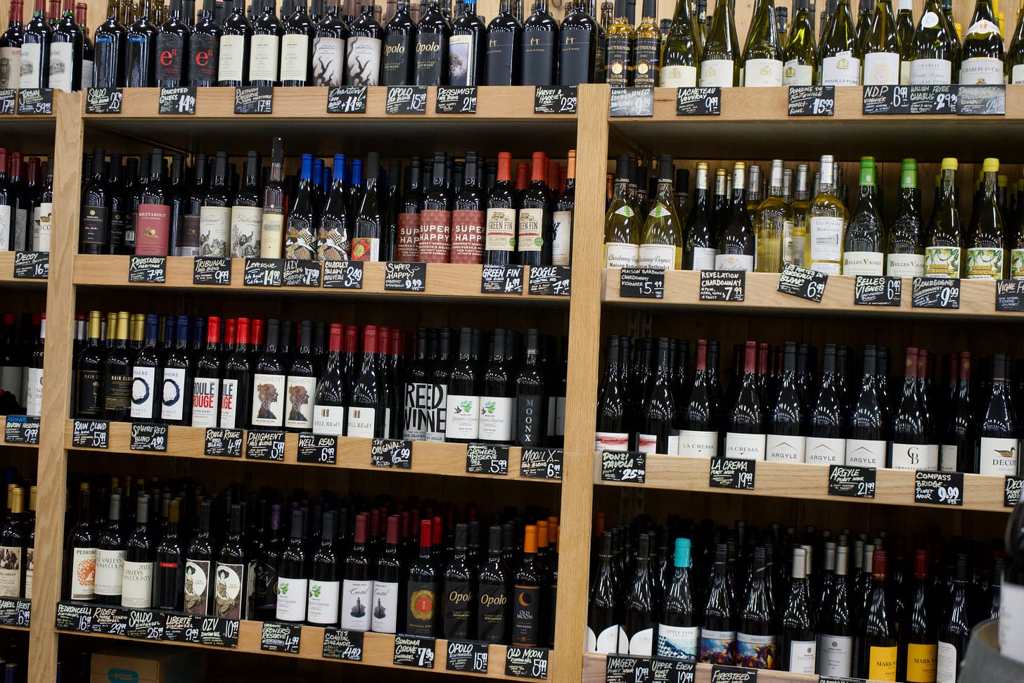 Wide view of wooden wine shelves filled with tightly arranged bottles, labels and chalkboard price tags forming repeating rows across the frame. The composition emphasizes pattern, abundance, and the quiet rhythm of everyday spaces.