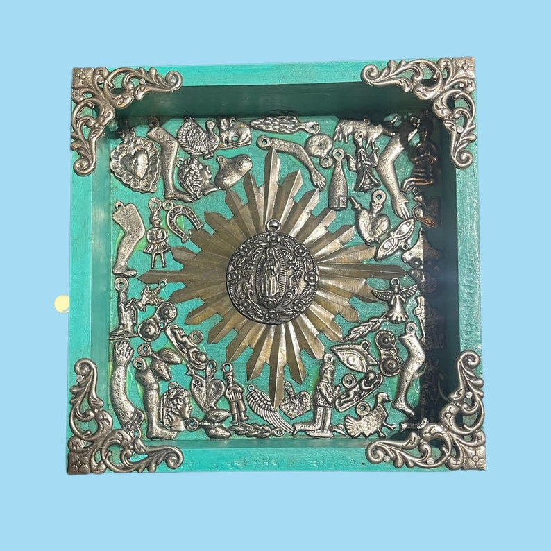 May include: A turquoise wooden tray with ornate metal corners. The tray is filled with a variety of silver charms, including hearts, hands, and a religious icon. The center of the tray features a gold sunburst design. May include: A turquoise wooden tray with ornate metal corners. The tray is filled with a variety of silver charms, including hearts, hands, and a religious icon. The center of the tray features a gold sunburst design.
