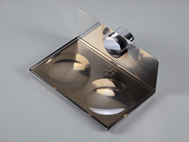 May include: A chrome soap dish with two compartments. The dish is mounted to a wall with a chrome screw. The dish has a clear glass back panel.