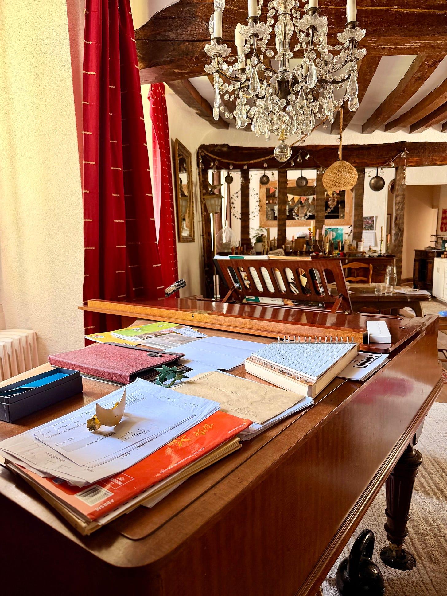 Stacks of paperwork, folders, notebooks and a music book covering the closed lid of a wooden piano in a French sitting room, with red curtains and a dining room beyond. Stacks of paperwork, folders, notebooks and a music book covering the closed lid of a wooden piano in a French sitting room, with red curtains and a dining room beyond.