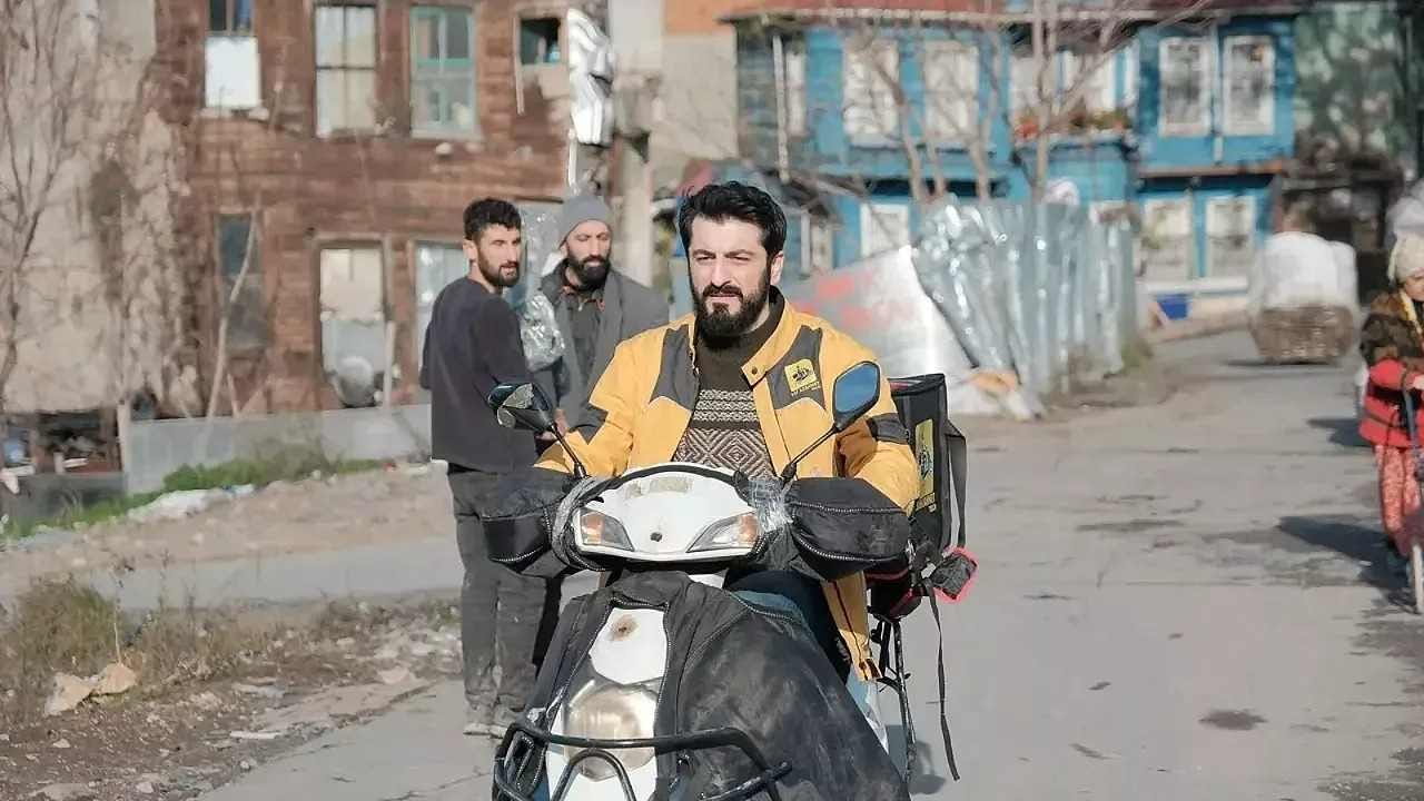 Sarı kuryeci montu giymiş Mahsun J karakteri, motosiklet üzerinde şehir sokaklarında ilerliyor; arka planda eski binalar ve mahalle sakinleri görülüyor. Sarı kuryeci montu giymiş Mahsun J karakteri, motosiklet üzerinde şehir sokaklarında ilerliyor; arka planda eski binalar ve mahalle sakinleri görülüyor.