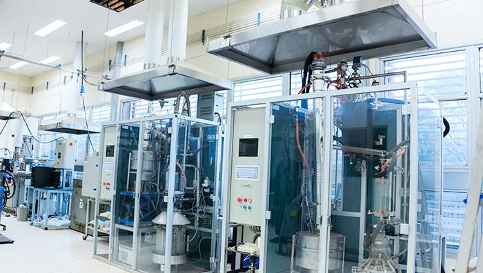 Interior de laboratório ou instalação industrial com múltiplas máquinas de grande porte encapsuladas em estruturas de vidro e metal branco, equipadas com painéis de controle, iluminação superior em painéis de teto, piso branco, uma pessoa em pé próxima a uma das máquinas à direita, equipamentos auxiliares e tubulações visíveis nas laterais, e janelas com venezianas azuis ao fundo. Interior de laboratório ou instalação industrial com múltiplas máquinas de grande porte encapsuladas em estruturas de vidro e metal branco, equipadas com painéis de controle, iluminação superior em painéis de teto, piso branco, uma pessoa em pé próxima a uma das máquinas à direita, equipamentos auxiliares e tubulações visíveis nas laterais, e janelas com venezianas azuis ao fundo.