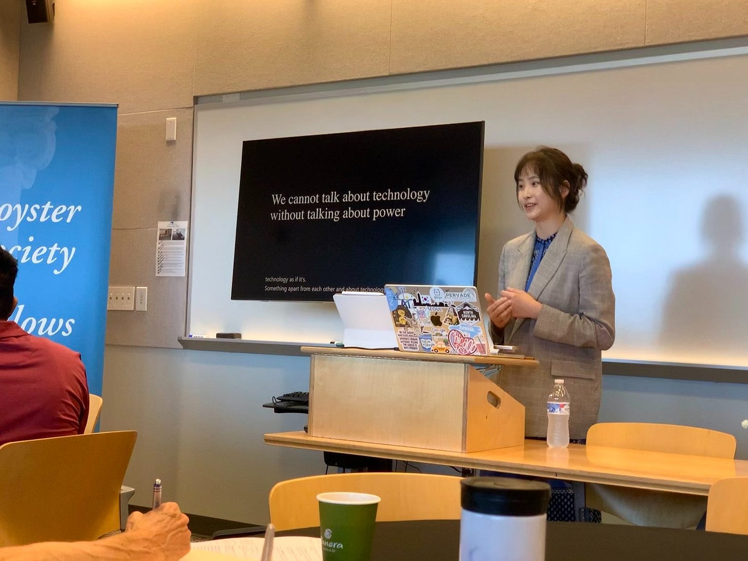 Heesoo Jang stands at a podium next to a display that reads "We cannot talk about technology without talking about power." In front of her is a laptop covered in stickers, and she wears a boxy blazer.