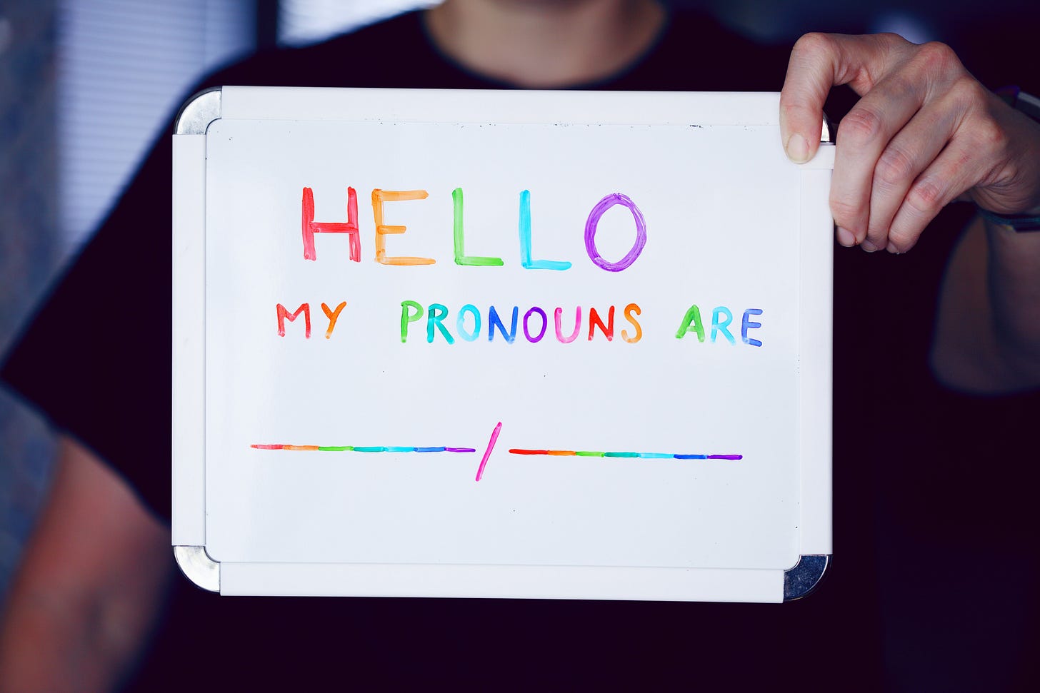 Person holding a white board with the following text written in rainbow colors: "Hello my pronouns are __/__." Person holding a white board with the following text written in rainbow colors: "Hello my pronouns are __/__."