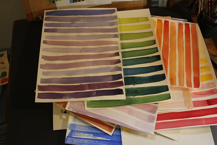 Watercolor practice sheets with rows of color swatches in purple, green, orange, and red tones stacked on a desk. Close-up of watercolor practice sheets showing strips of purple, green, yellow, and red color swatches arranged diagonally.