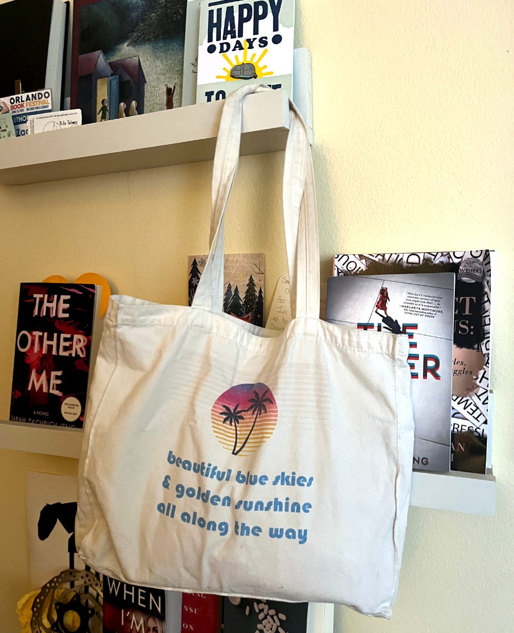 A tote bag hangs from a shelf full of various other bookish items. The bag has an ‘80s inspired design of palm trees and the catchphrase “Beautiful blue skies and golden sunshine all along the way” which David Lynch used to end his weather reports on KCRW radio. A tote bag hangs from a shelf full of various other bookish items. The bag has an ‘80s inspired design of palm trees and the catchphrase “Beautiful blue skies and golden sunshine all along the way” which David Lynch used to end his weather reports on KCRW radio.
