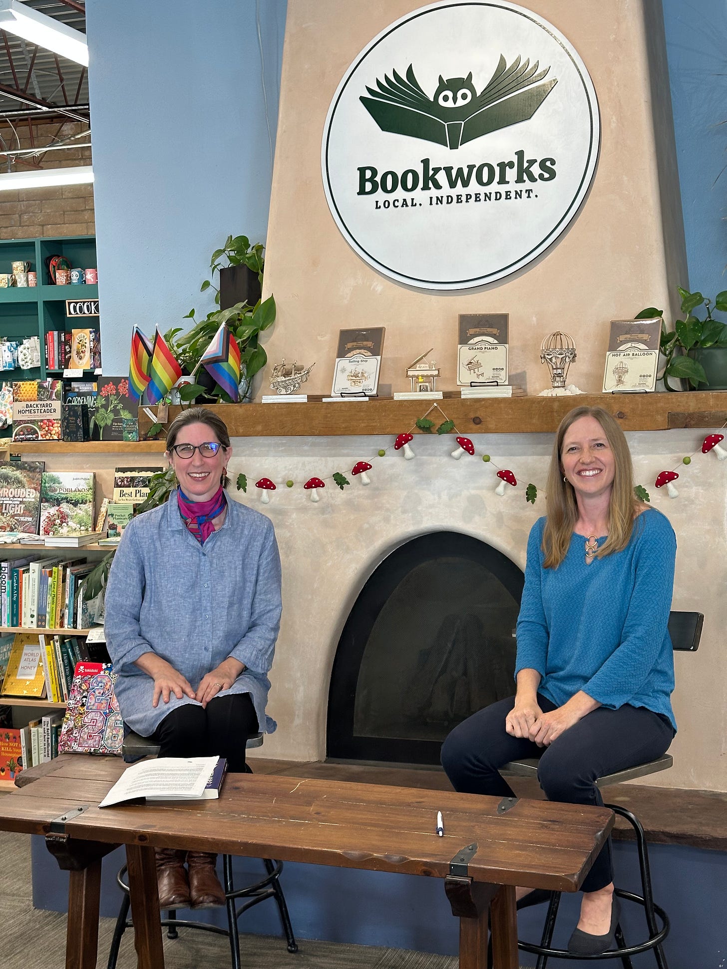 Me and Elise Nye sit in front of a kiva-style fireplace, both of us grinning and wearing blue tops. Several books are on the mantel and the Bookworks sign is on the wall in the background.