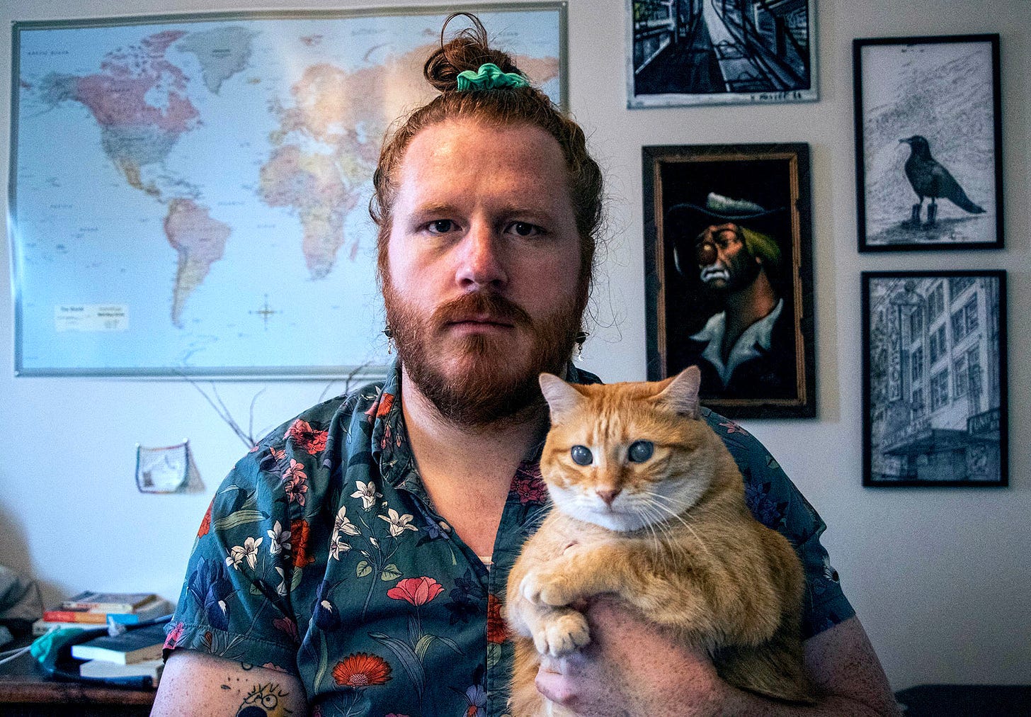 A bearded person wearing a floral button-up short-sleeve shirt, orange hair tied up in a high bun looks into the camera. They are holding an orange cat with cloudy eyes, which is also staring into the camera.