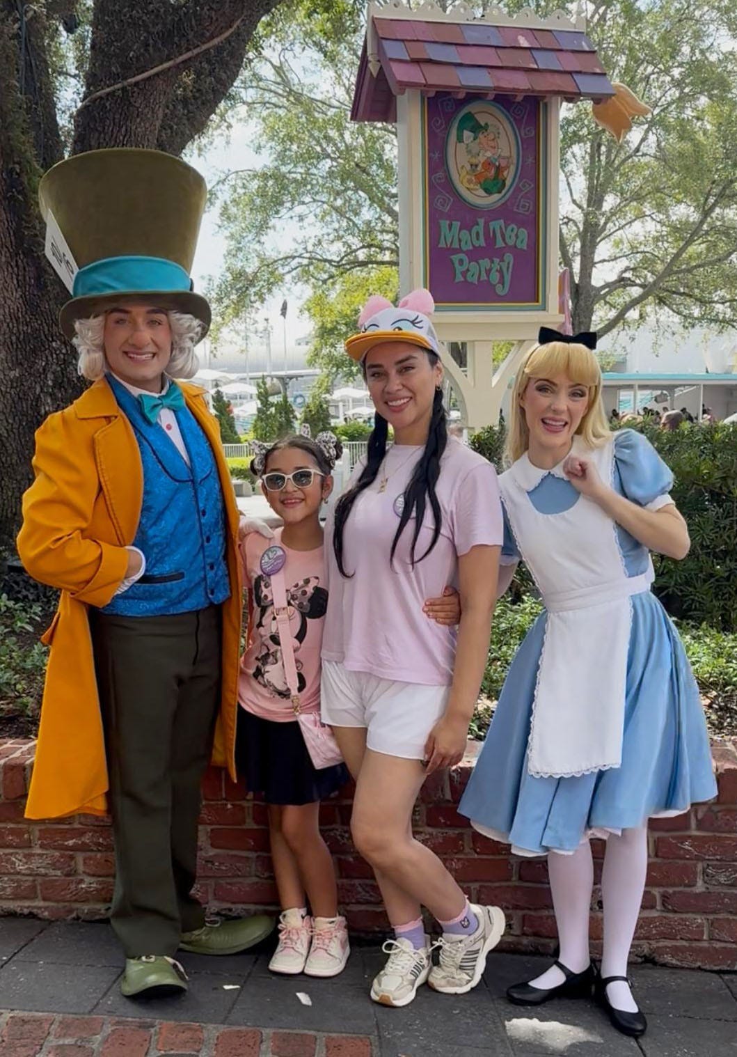 Four people smile and pose for a photo in front of a sign that says “Mad Tea Party”: one person is dressed as the Mad Hatter from “Alice in Wonderland,” one is dressed like Alice, one is a young girl wearing a Mickey Mouse shirt and one is a woman wearing a Daisy Duck visor. Four people smile and pose for a photo in front of a sign that says “Mad Tea Party”: one person is dressed as the Mad Hatter from “Alice in Wonderland,” one is dressed like Alice, one is a young girl wearing a Mickey Mouse shirt and one is a woman wearing a Daisy Duck visor.