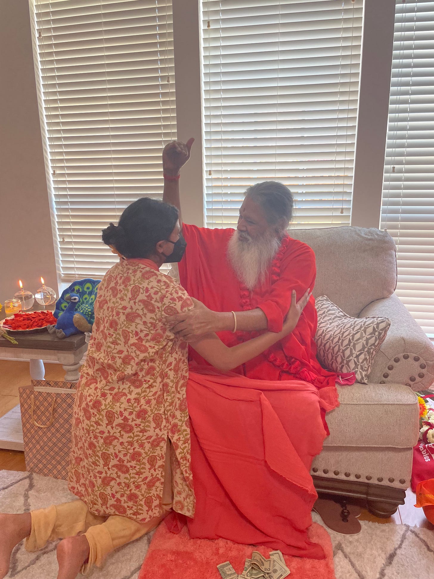 Vahni, an Indian woman in a floral salwar and black medical mask, embraces Sri Ganapathy Sachidananda Swamiji, an Indian guru in pink robes with a long white beard and smiling face