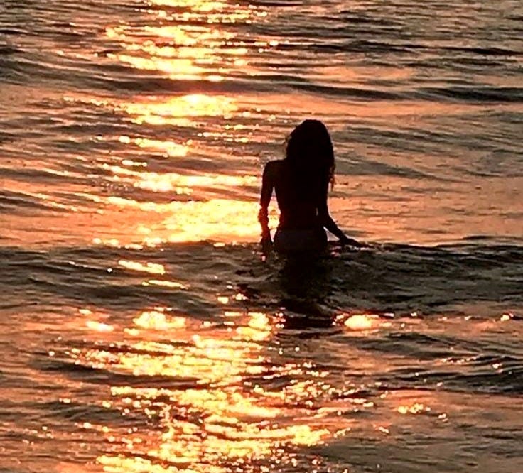 This may contain: a woman in the water at sunset with her back turned to the camera and sun reflecting on the water