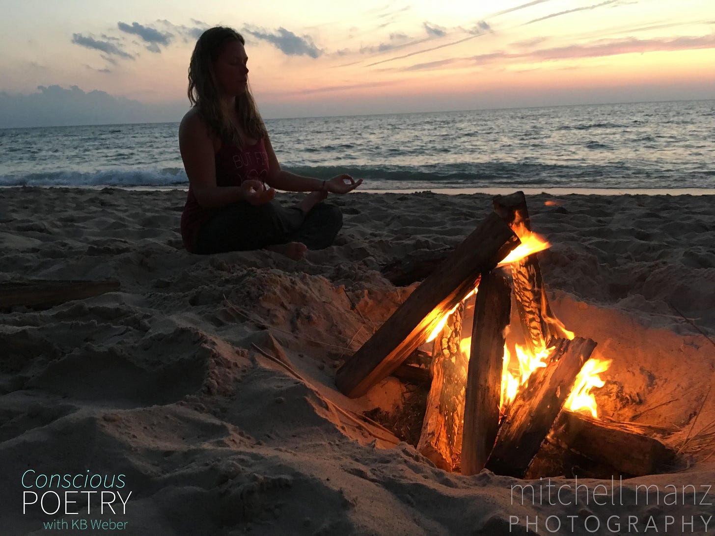 Woman sitting in the sand next to Lake Michigan during sunset with light blues, yellows and pinks. She sits in meditation next to a roaring fire dug into the sand