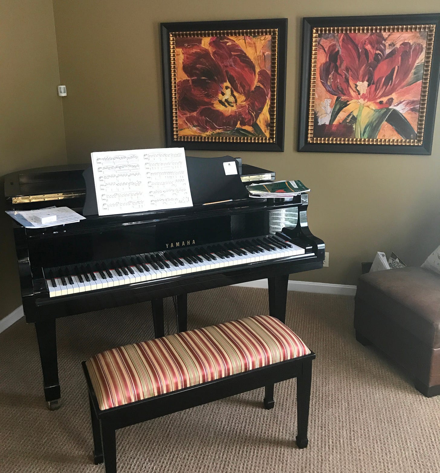 A cozy music room featuring a black Yamaha baby grand piano with sheet music on its stand. The piano is paired with a striped, upholstered bench. Behind the piano are two framed, vibrant floral paintings, creating a warm and artistic ambiance. The room is carpeted and has a comfortable seating area visible in the corner, emphasizing a lived-in and inviting space for musical practice and creativity.
