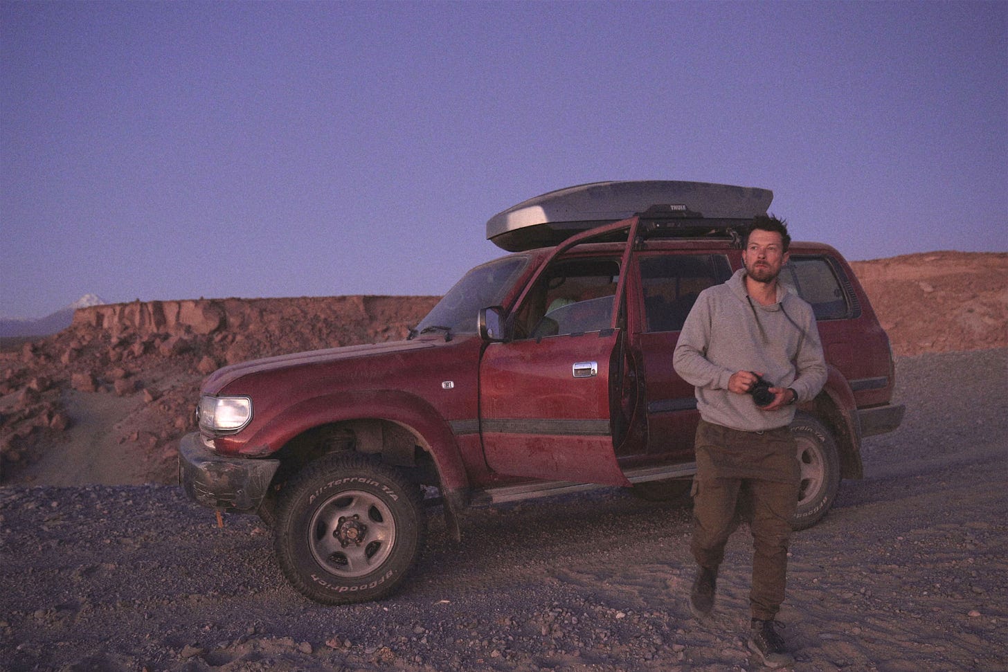 A man holding a camera walks away from a red SUV with an open door, parked on a rocky landscape under a purple sky at dusk.