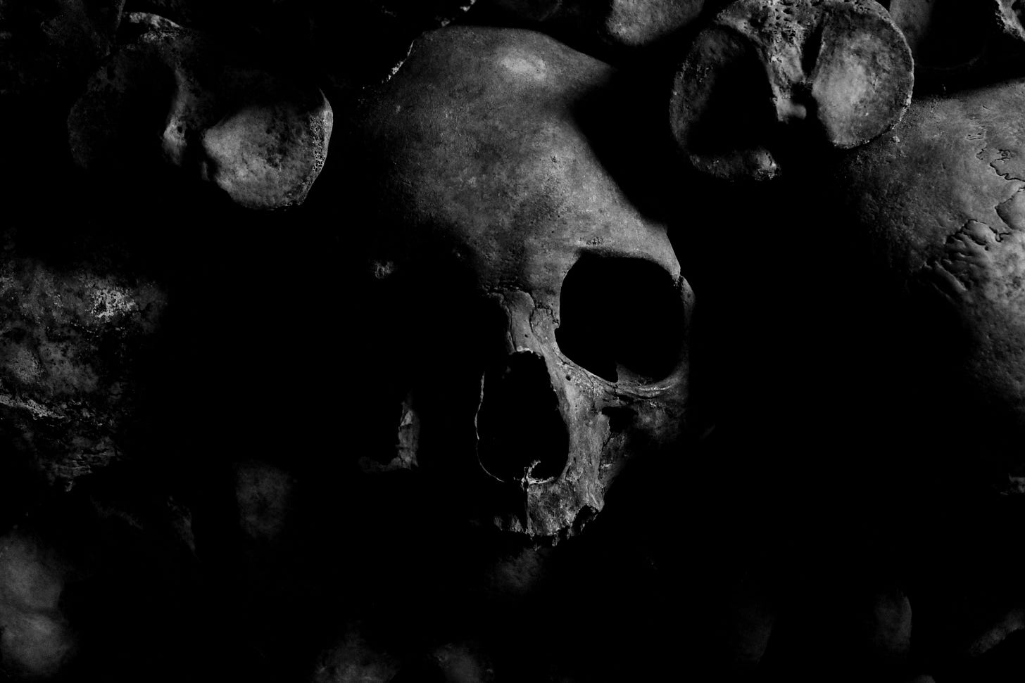 A black and white image centered on a human skull, surrounded by other skulls which are obscured in shadow.