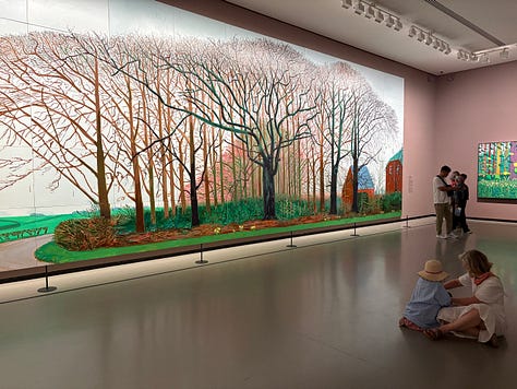 Multiple bright-colored paintings including a self-portrait of Hockney in a yellow plaid suit and big yellow glasses, the crowd viewing paintings, a mother and child in front of a bright painting of a massive tree, an annunciation painting redone in neon colors, a green lawn with sprinkler spray, a photograph of hockney working at his drawing table with his little terrier dog under the table and two paintings with raindrops hitting puddles among trees