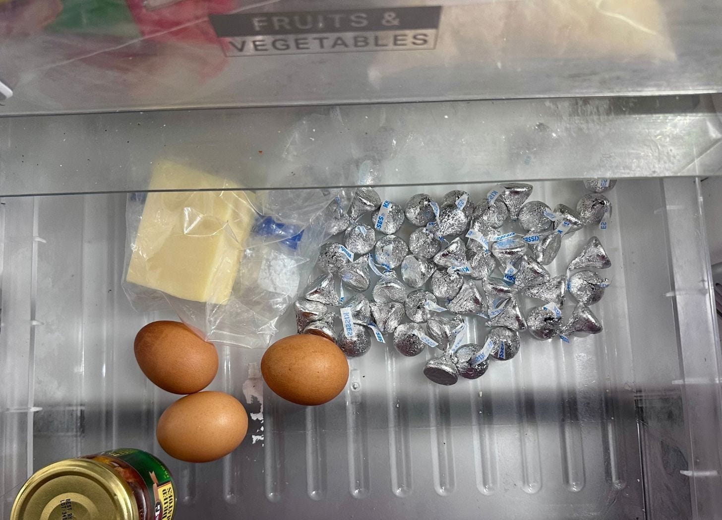 An open refrigerator vegetable drawer with a few dozen foil-wrapped Hershey's Kisses spilled across one end, three brown eggs, a block of white cheese in a plastic bag, and a jar of sun-dried tomatoes. The drawer label reads Fruits and Vegetables.