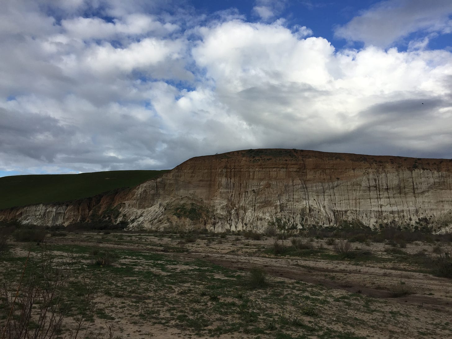 the riverbed, dry and lightly vegetated, stretches out ahead. on the horizon the sand-coloured cliffside gently slopes and a hint of green hillside is visible beyond it. the sky is blue with lots of heavy white clouds.