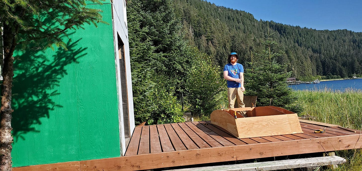 A photo of a smiling redheaded man posing with an upside down partially assembled boat hull in the sunshine