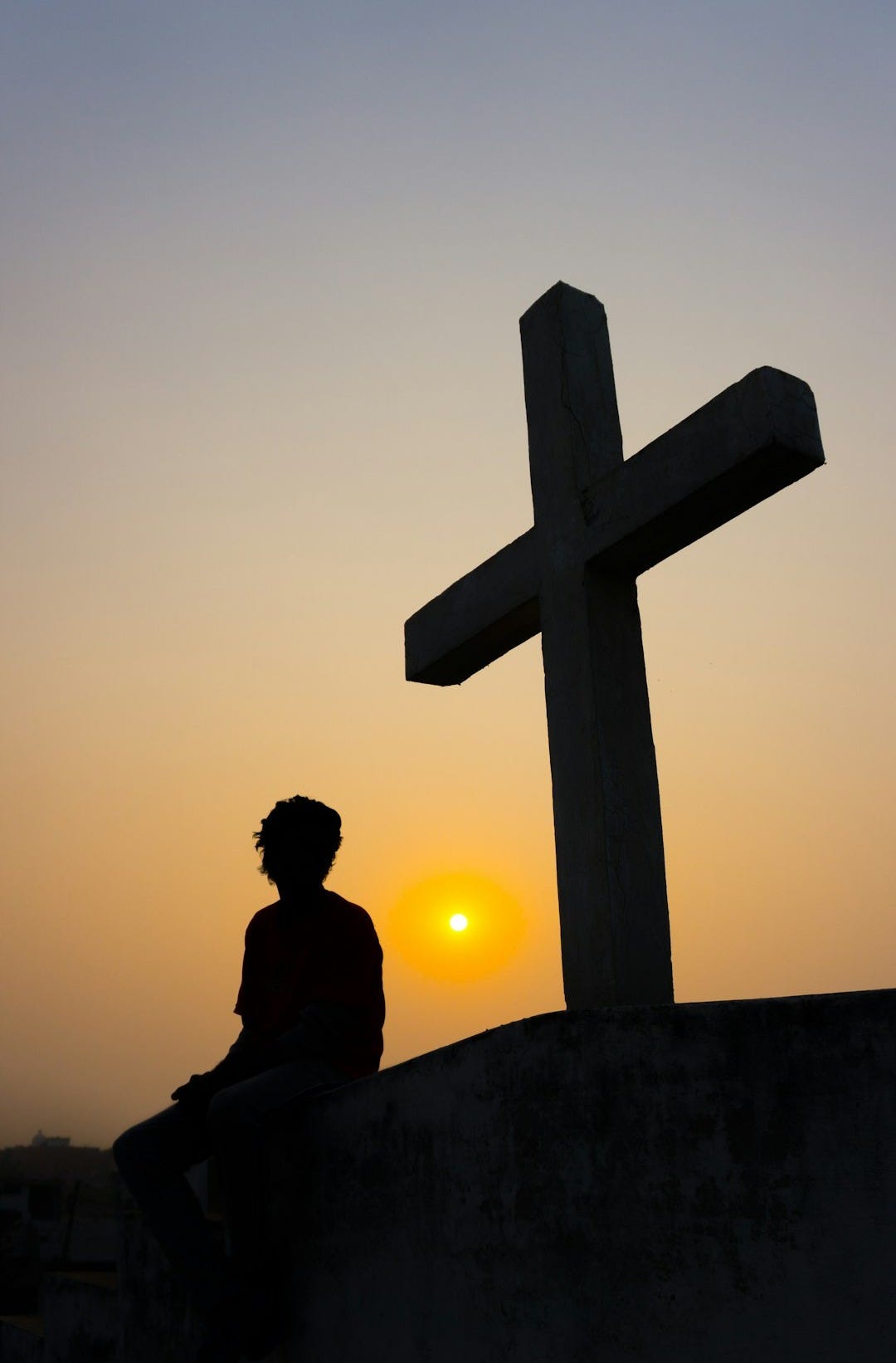 silhouette of man sitting on the edge of a cross during sunset
