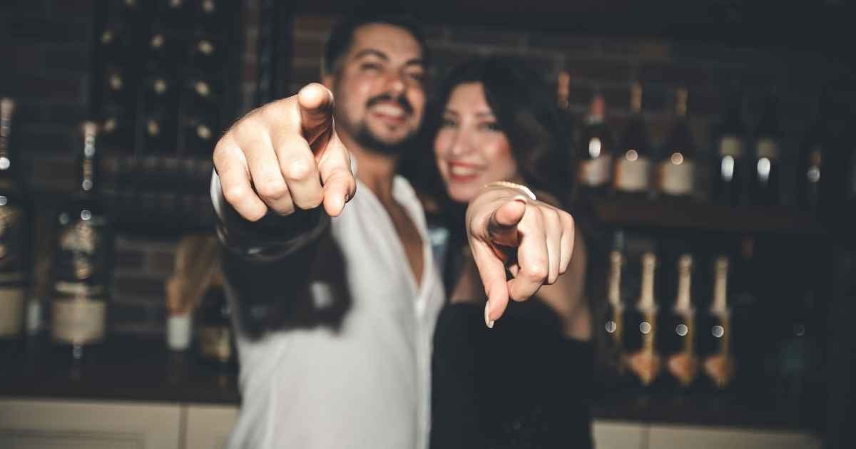 Man and woman pointing playfully towards the camera inside a cozy bar setting Man and woman pointing playfully towards the camera inside a cozy bar setting