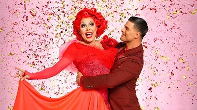 La Voix wears a bright red ballgown and holds the skirt in one hand, posing in a lift with Aljaz, who wears a dark red suit.