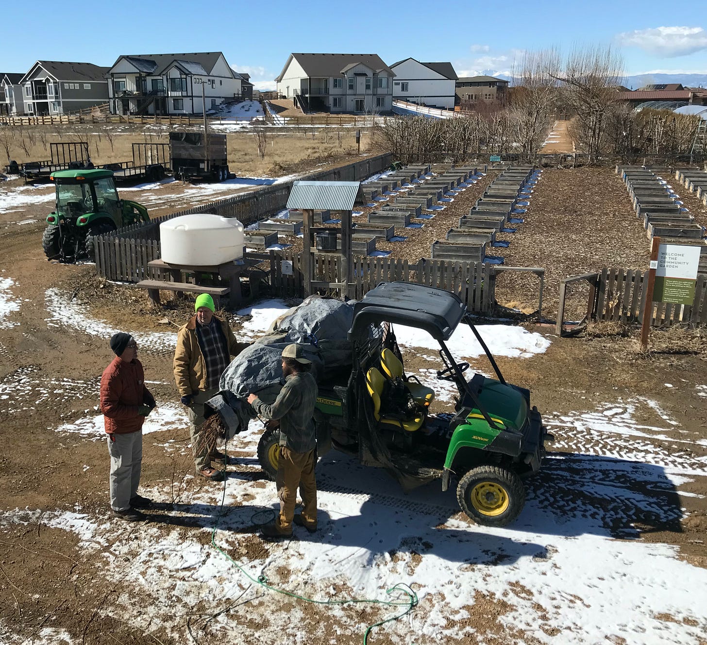 Parked ATV with a bundle of willow on the back. Three men standing and talking at the back of the ATV. A community garden, willow trees, and a suburban housing development in the background. 