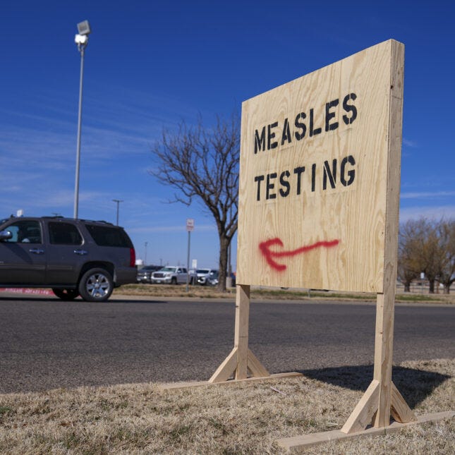 A wooden sign spray-painted with the black letters "MEASLES TESTING" and a red left-pointing arrow below stands on the side of a road as a car passes -- Health coverage from STAT