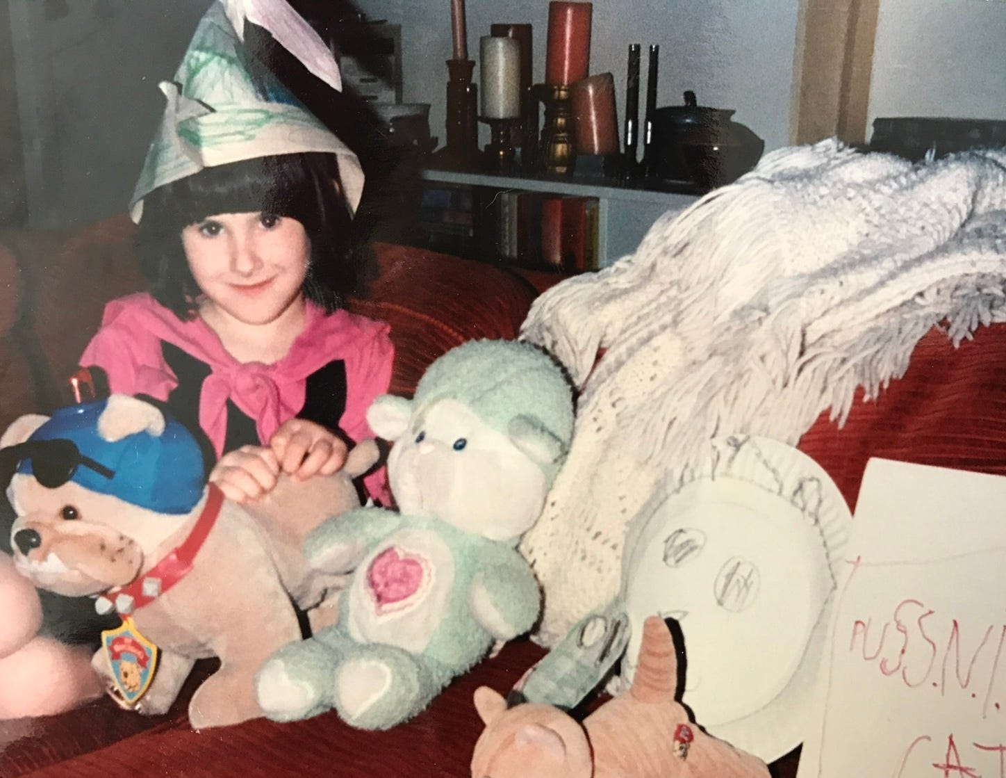 Photo of a young Lyric, sitting on a red sofa with a white throw, dressed in a costume they made themselves that's supposed to be Puss N Boots. Several of Lyric's favorite stuffed toys are lined up on the sofa beside them.
