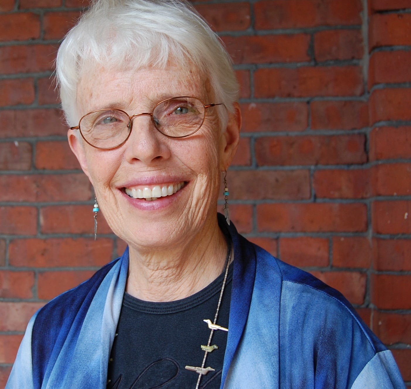 An elderly woman with white hair and glasses, a blue gown and a necklace with birds