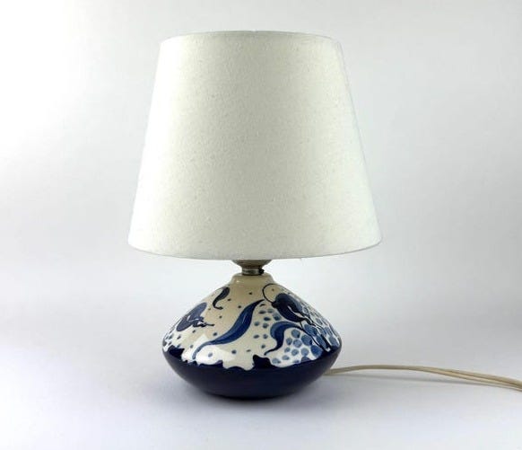 May include: A ceramic table lamp with a white lampshade. The base is dark blue with a painted design in white and blue. The design appears to be floral or abstract. A cream-colored cord extends from the base.