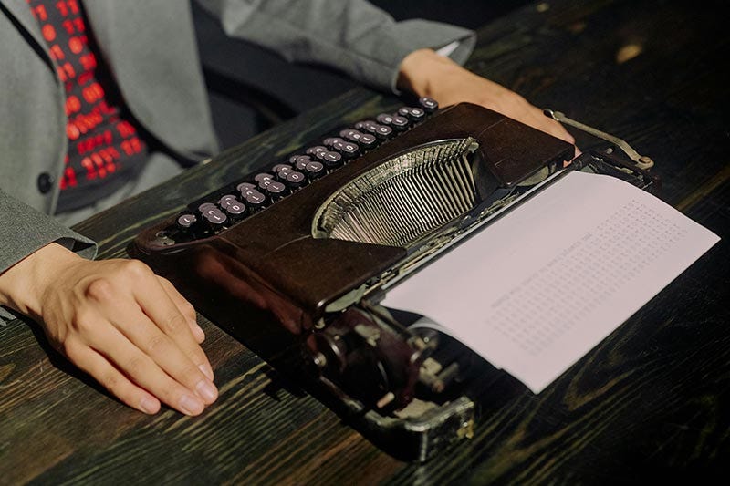 Une personne a les mains posées sur son bureau, encadrant une machine à écrire de laquelle pendent des feuilles quasi-vierges.