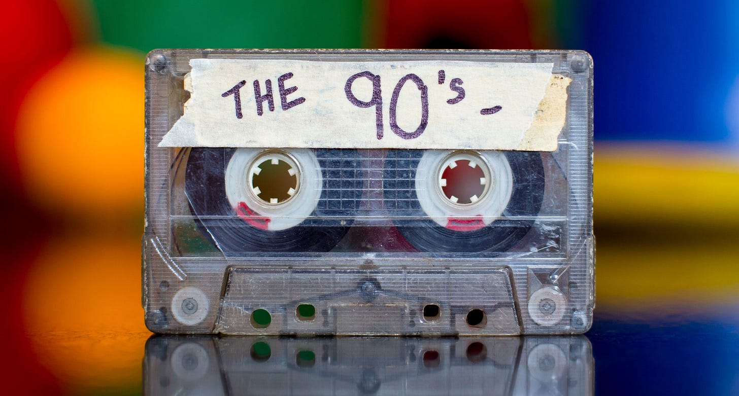 Best 90s Songs - A photograph of a scratched and beat up cassette tape that's got. a piece of tape adhered to the front with the label, 'The 90s,' on it. It sits on a reflective surface and there's a colorful blurred background behind it.