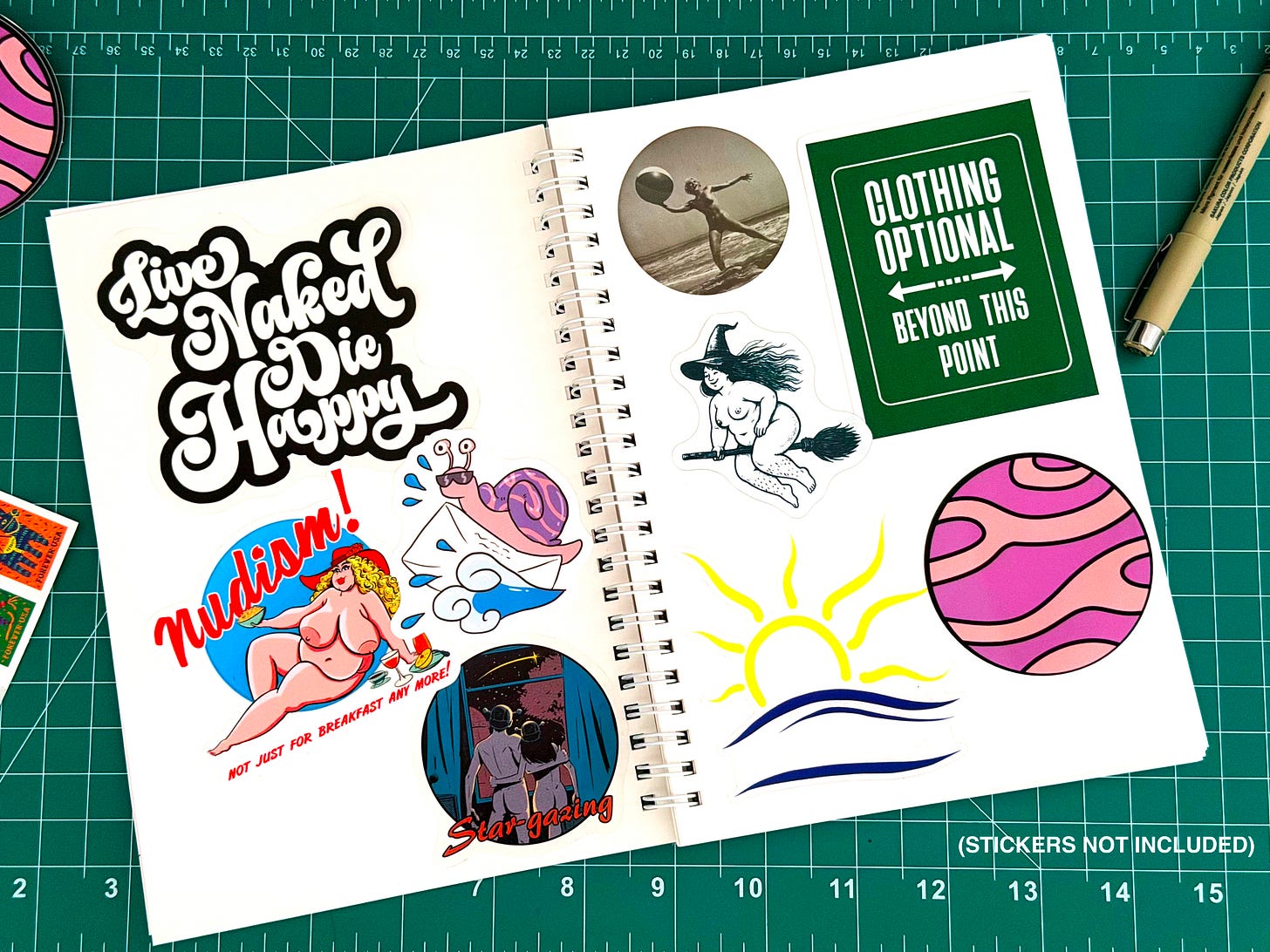 The Planet Nude Sticker Book open on a green cutting mat, pages filled with nudist-themed stickers including "Live Naked Die Happy," a nude witch on a broomstick, a "Clothing Optional" sign, the Planet Nude snail mascot, and others. A note in the corner reads: stickers not included.
