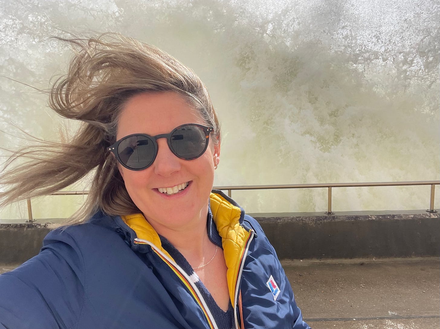 A woman with brown hair and blue jacket in front of a giant wave