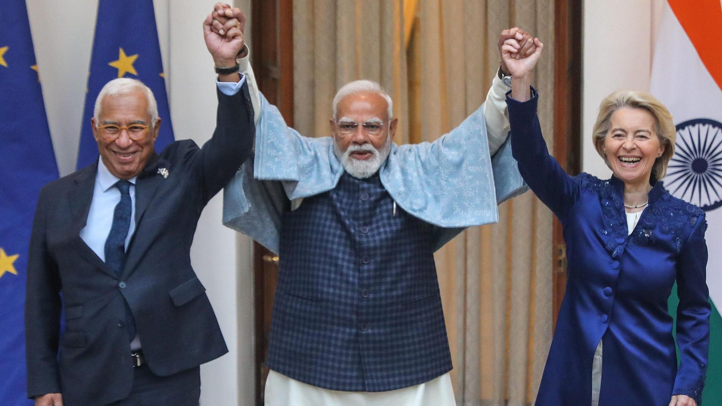 Three leaders hold hands and raise their arms, laughing during a friendly moment. The man in the centre, Indian Prime Minister Narendra Modi, with a white beard and glasses, wears a light shawl over a dark outfit. On either side, European Council President António Costa in a dark suit and European Commission President Ursula von der Leyen in a dark blue long jacket