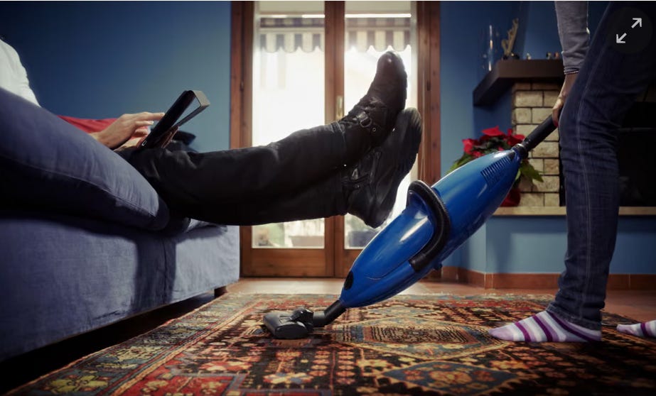 Stock image of a close-up of a hoover that a woman is using on a carpet by a sofa. A man is on the phone on the sofa with his feet up.