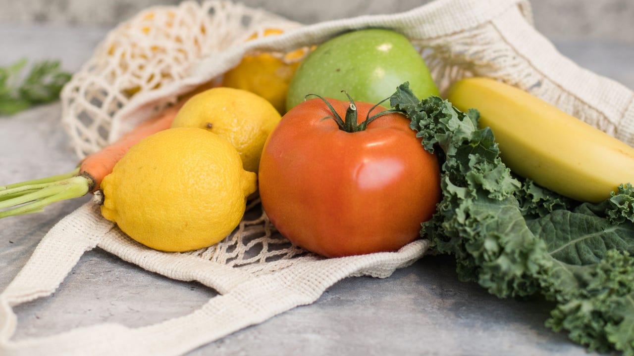 A cloth produce bag filled with fresh fruits and vegetables, including tomatoes, lemons, a green apple, a banana, a carrot, and kale on a light gray surface