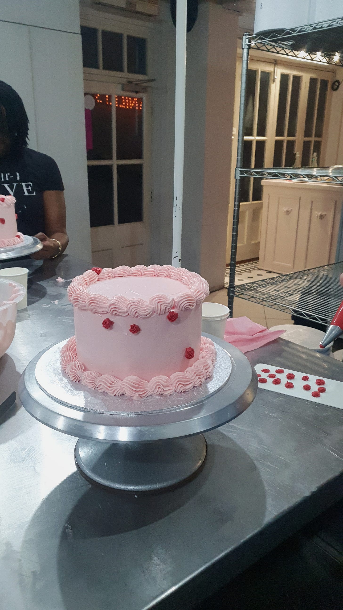 Photo of a pink cake with a few red roses piped on and a scraper to the side with lots of practice iced roses on it