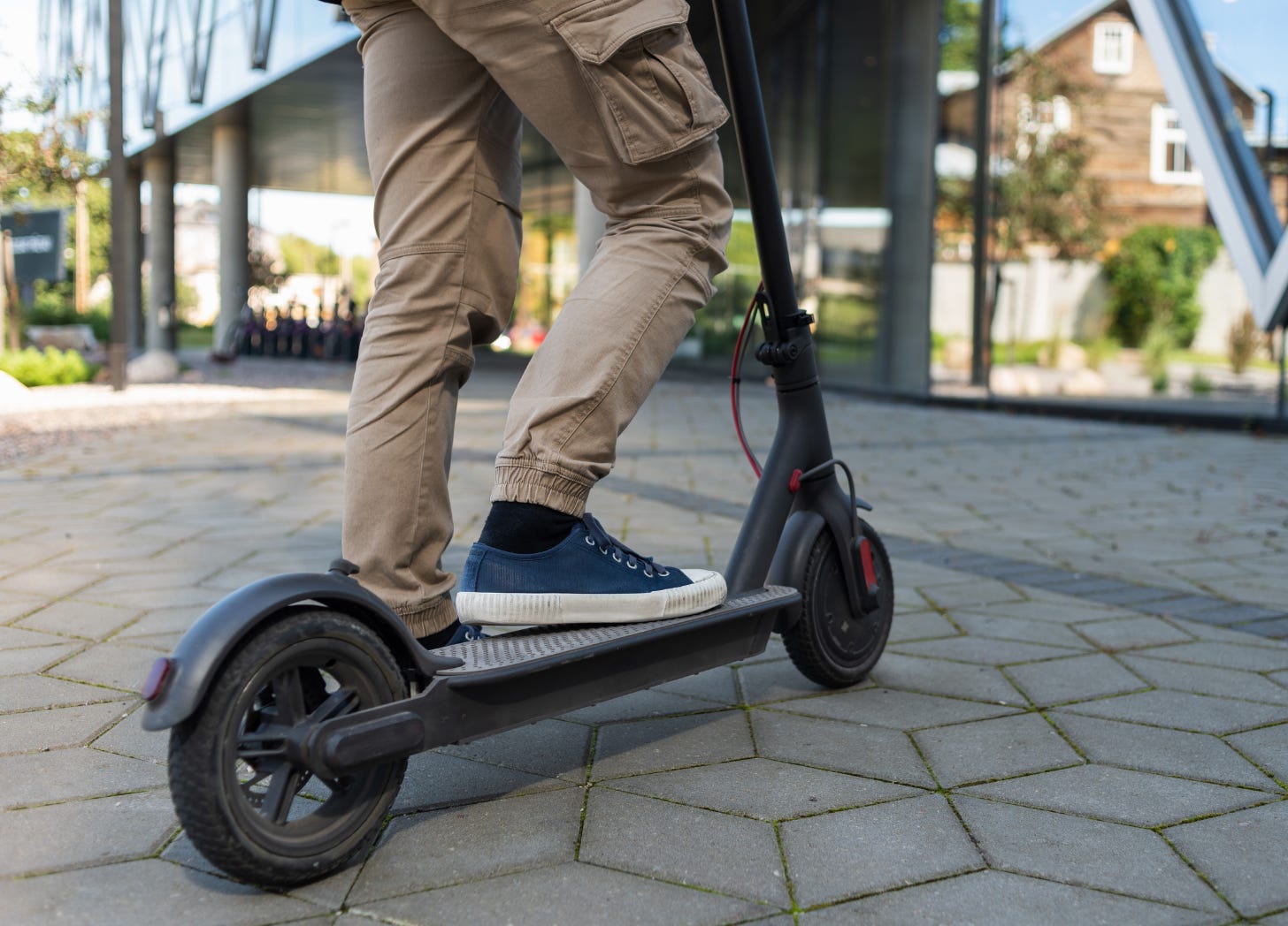 Young child wearing a backpack rides an electric scooter on pavement with houses in the background.