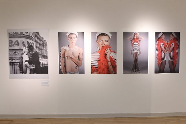 1) Gallery wall with five portraits. The first is a black-and-white image of a two people kissing. It's the final frame of Tessa Boffin's The Sailor and the Showgirl, where they are kissing in front of a Sanyo building. The others show a person in sailor attire and an orange feather boa, exuding confidence. There are 4 modern-day images of the sailor with the boa, the first one, the sailor is shirtless with a heart nipple pastie on. The second image, the sailor is clothed with a striped shirt, and the orange boa. The third image is a full-body image of them holding the sides of their head, and the final image is a mirror image of the sailor holding hands with themself. There is a placard beneath Tessa's work that gives some context about her piece, and the inspired student's piece next to it. 2) Young white skinny person wearing a white sailor hat, holding a white and blue/black striped shirt, with various tattoos including an anchor, ribbon, and barb wire, and HIV AIDS. The expression is neutral. There is a red lipstick print on their neck. They are shirtless, with a red heart nipple pastie over their right nipple. They have short brown hair, and they are staring straight into the camera. They are standing in front of a grey backdrop. They are holding the shirt over their right shoulder.