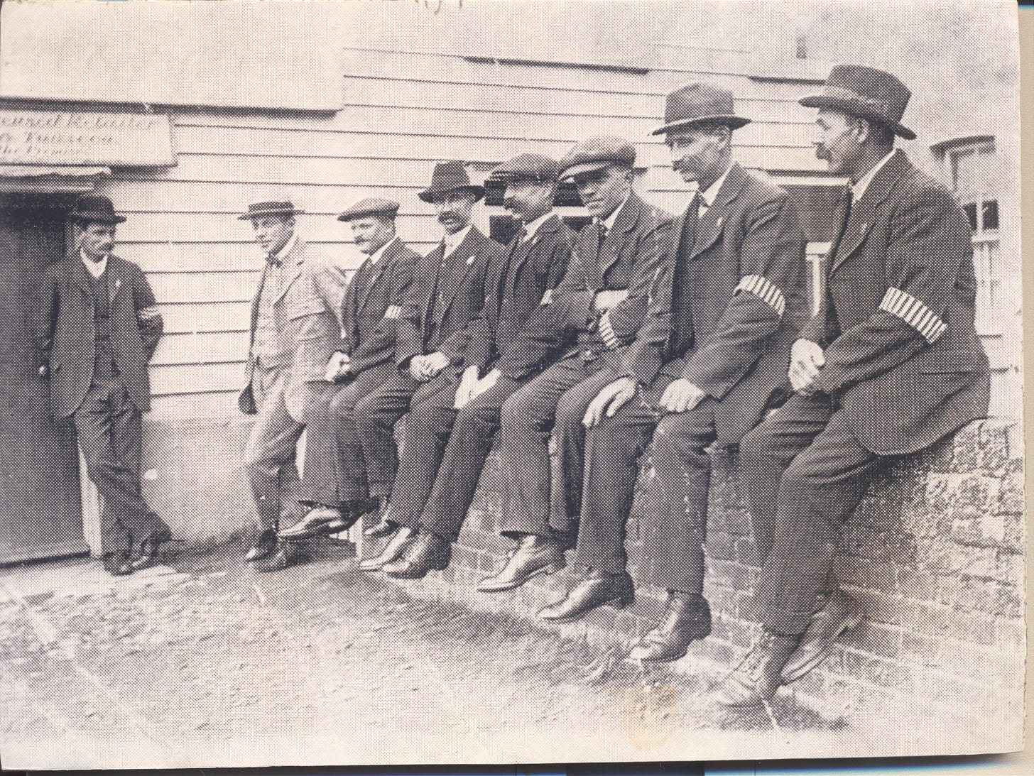 7 men sitting on a low wall wearing suits and a striped Special Constabulary armband. To the left another man, Edgar Nicholas, is standing leaning against the building.