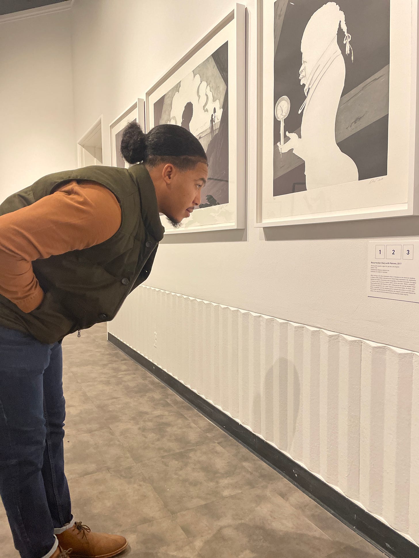 A man dressed in jeans, a brown shirt, and a green vesting, leaning over to read a small wall panel in front of three black and white art prints A man dressed in jeans, a brown shirt, and a green vesting, leaning over to read a small wall panel in front of three black and white art prints