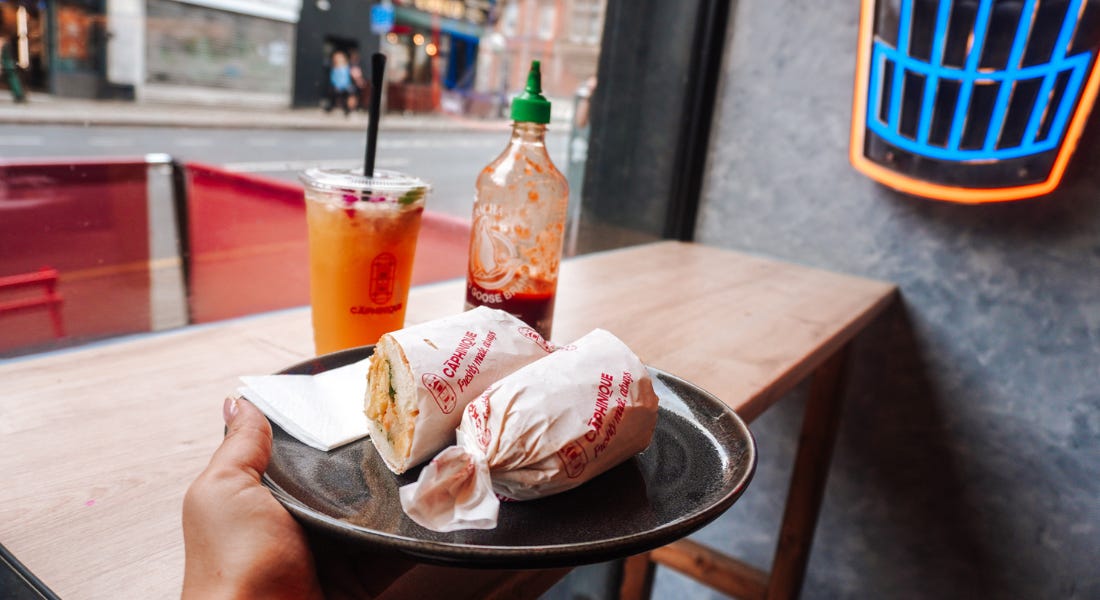 A hand holding a brown plate with a roll on it, with a fruit drink and sriracha bottle behind it