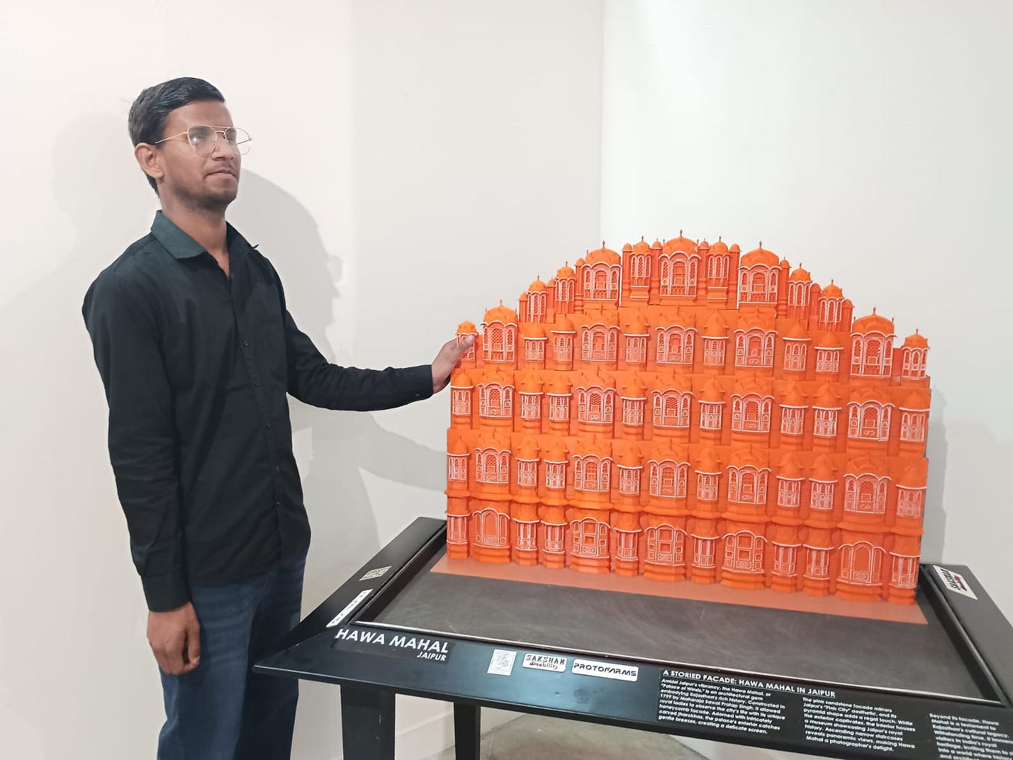 A man wearing glasses and a black shirt with blue jeans stands beside a detailed orange model of Hawa Mahal displayed on a black exhibition table. He rests one hand on the model, which features the palace’s distinctive multi-tiered façade with numerous small arched windows, against a plain white wall background. A man wearing glasses and a black shirt with blue jeans stands beside a detailed orange model of Hawa Mahal displayed on a black exhibition table. He rests one hand on the model, which features the palace’s distinctive multi-tiered façade with numerous small arched windows, against a plain white wall background.